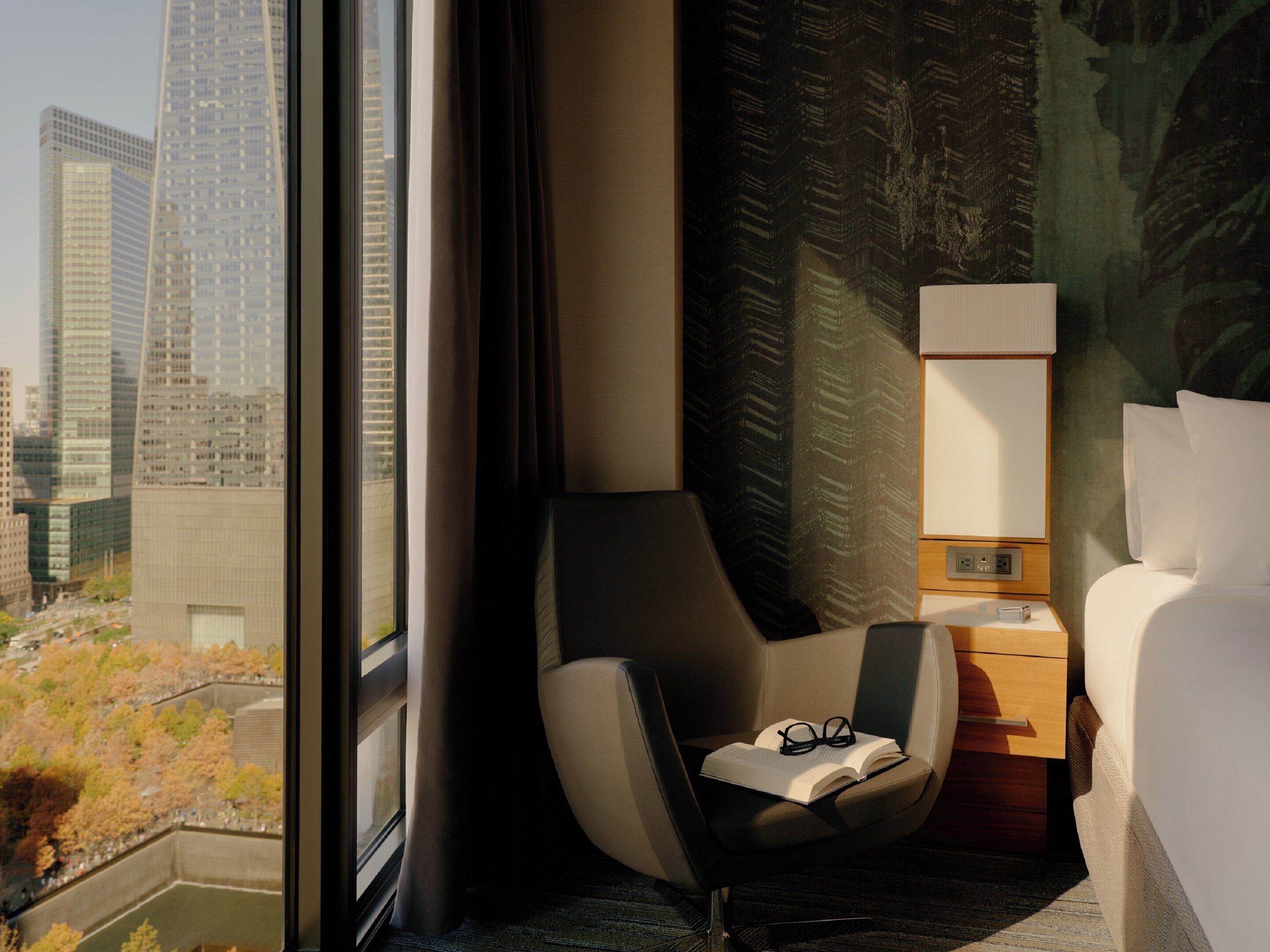 Quarto de hotel com vista para o horizonte urbano, uma cadeira moderna, mesa de cabeceira com abajur e uma cama bem arrumada.