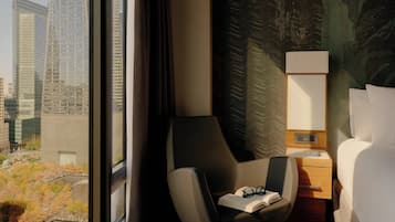 Una habitación de hotel con vistas a un paisaje urbano, una silla moderna, una mesita de noche con lámpara y una cama bien hecha.