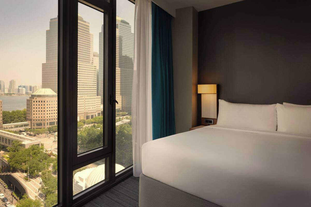 Een hotelkamer met een groot raam dat uitzicht biedt op de stad, een opgemaakt bed en een nachtkastje met een lamp.