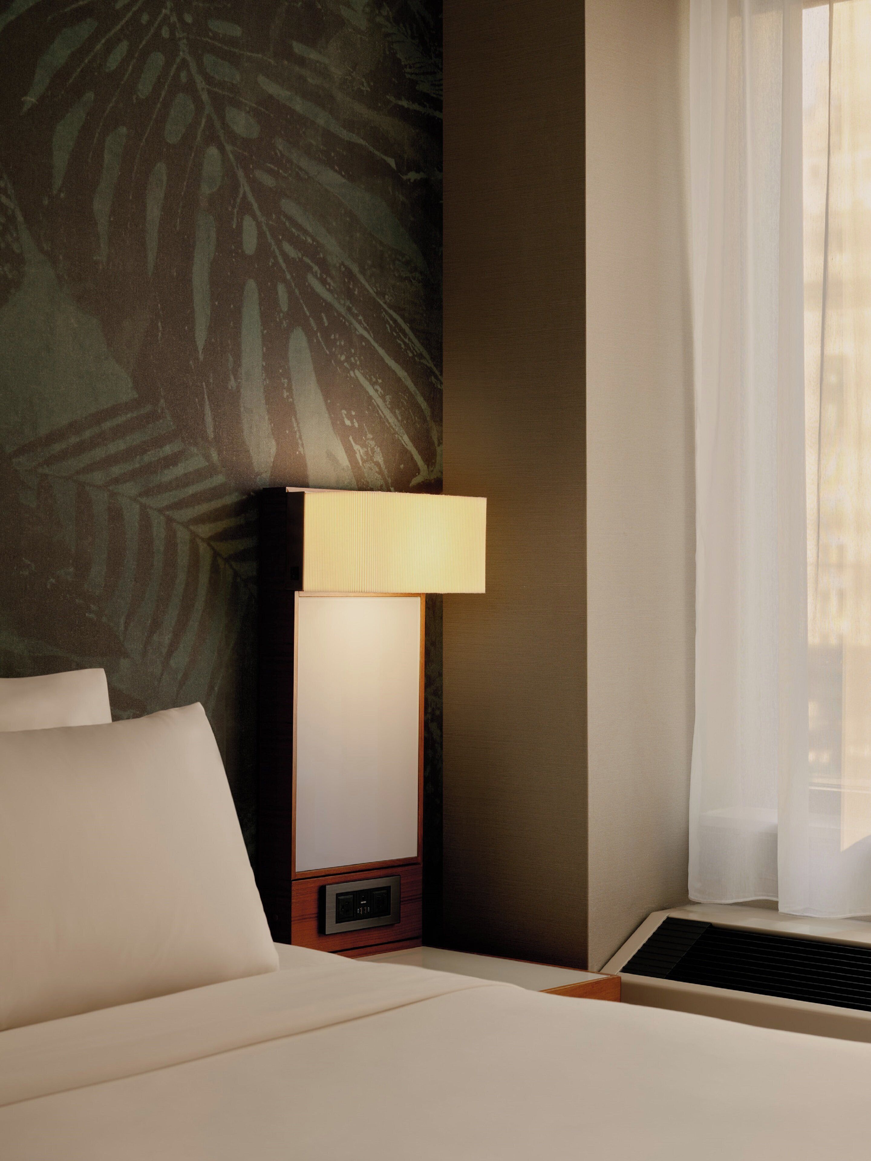 A hotel room with a bed, a nightstand with a lamp, a wall with a leaf pattern, and a window with sheer curtains.