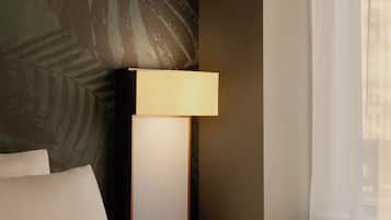 A hotel room with a bed, a nightstand with a lamp, a wall with a leaf pattern, and a window with sheer curtains.