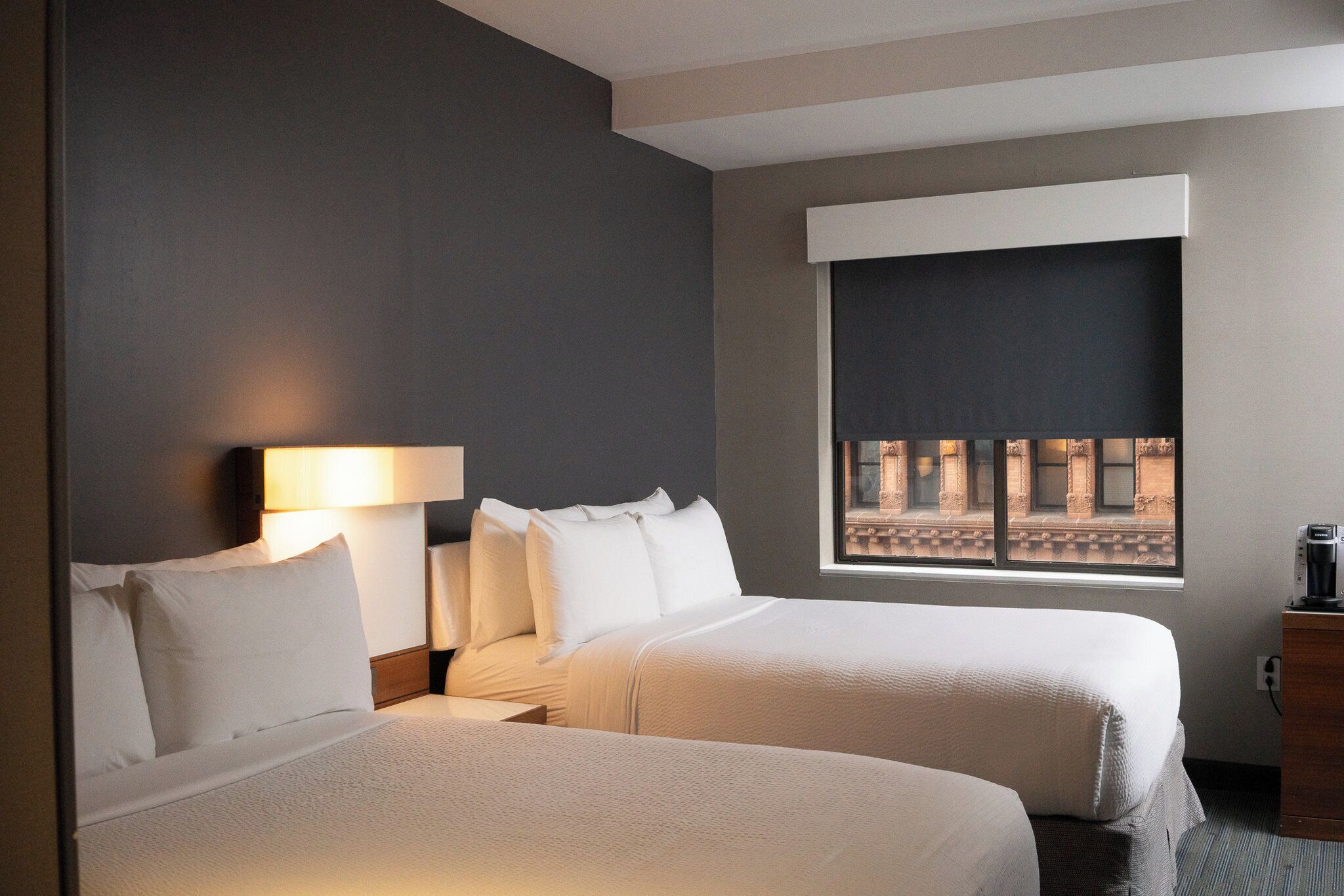 A hotel room with two beds, a nightstand with a lamp, a window with a view of buildings, and a wooden dresser.