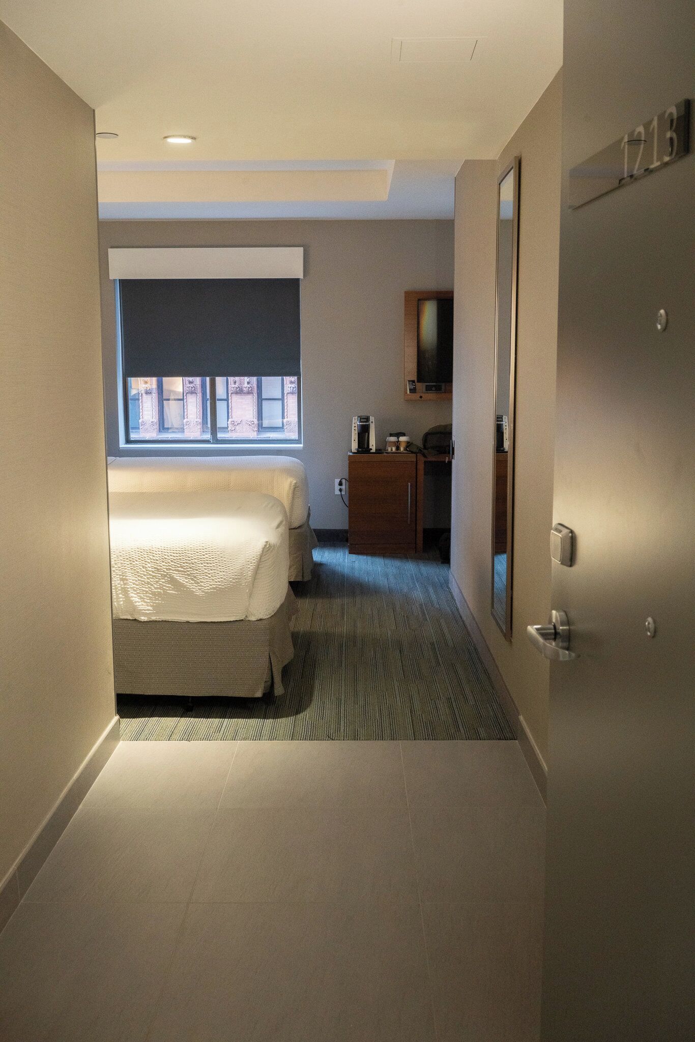 A hotel room with a bed, a small desk, a mirror, and a window.