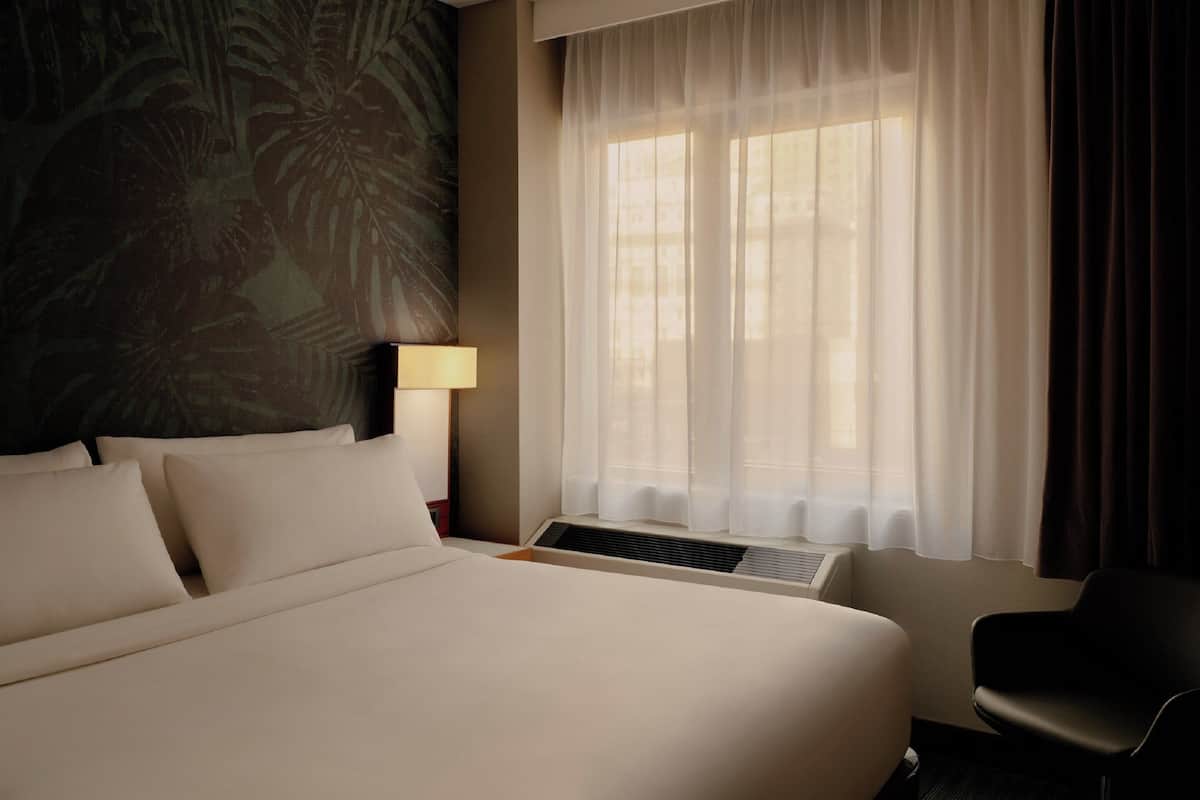 A hotel room with a large bed, a desk, a chair, a window with sheer curtains, and a wall with a tropical leaf pattern.