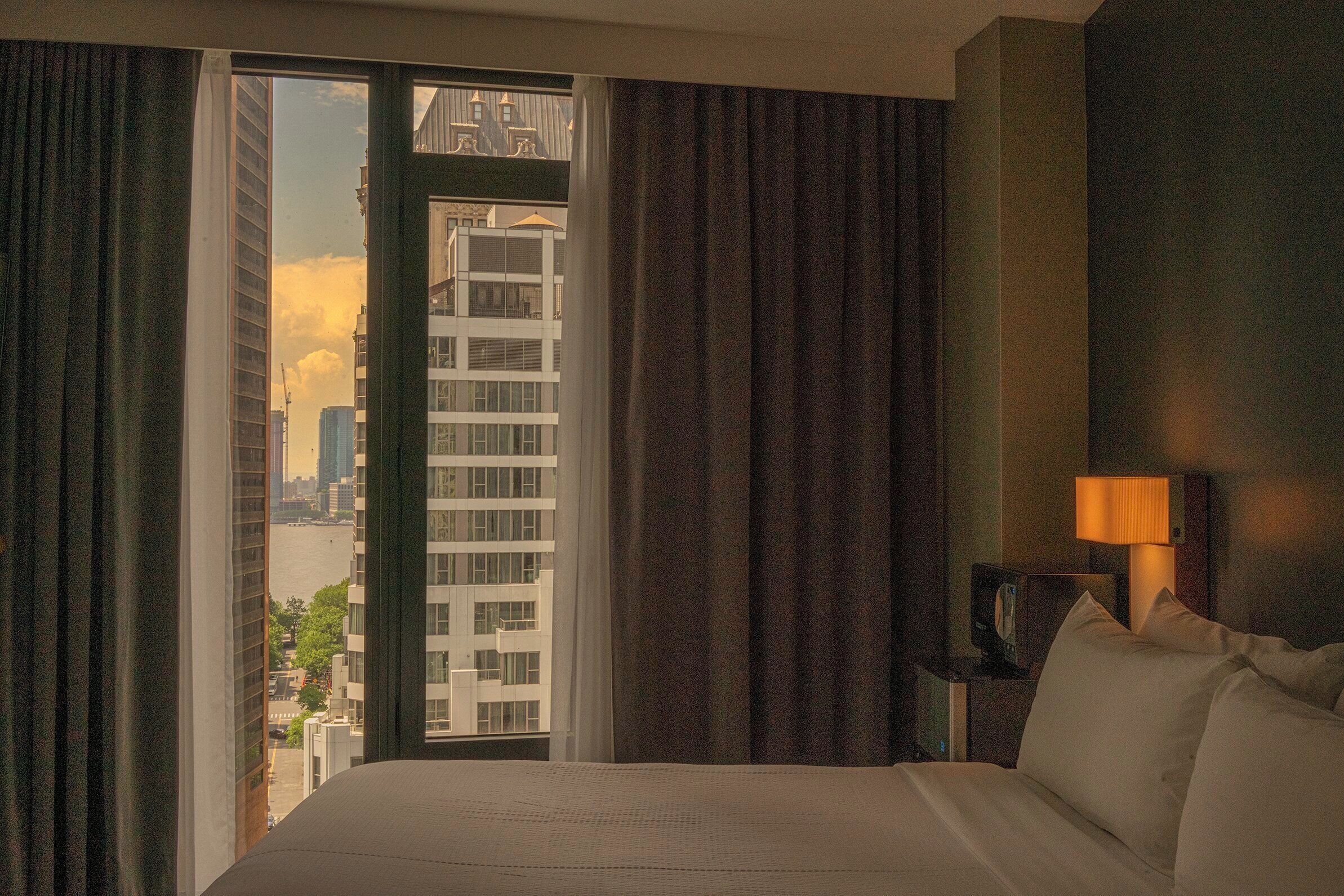 A hotel room with a large bed, a nightstand with a lamp, and a view of a cityscape through a window with curtains.