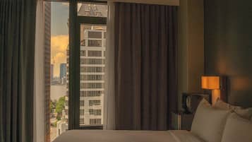 A hotel room with a large bed, a nightstand with a lamp, and a view of a cityscape through a window with curtains.