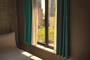 Habitación de hotel con un ventanal grande, una cama y vistas a edificios altos y árboles.