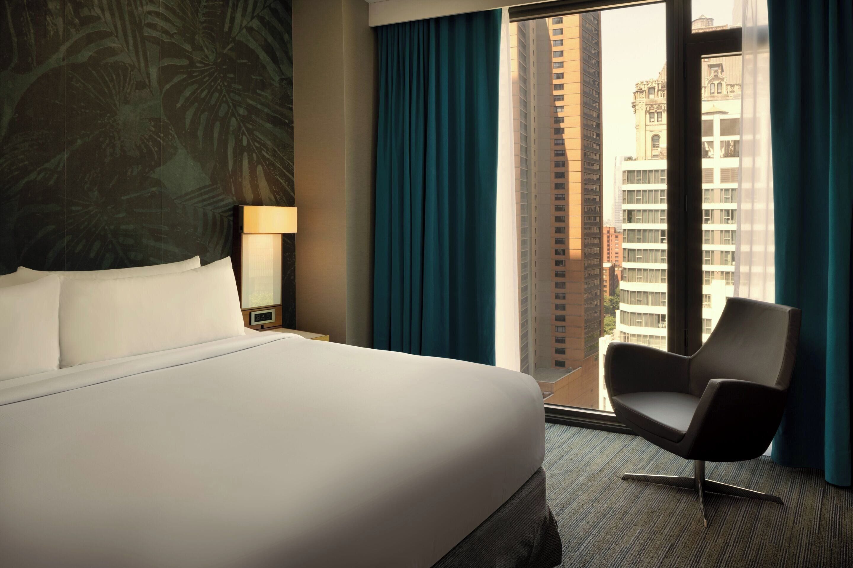 A hotel room with a large bed, a chair, a nightstand, a lamp, and a view of the city through a window.