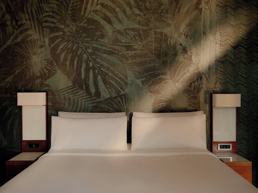 Una cama con dos almohadas, flanqueada por dos mesitas de noche con lámparas, frente a una pared con diseño de hojas tropicales.