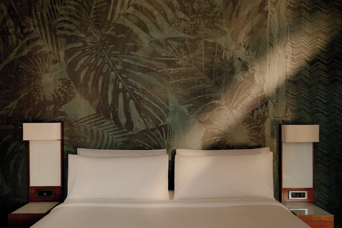 A bed with two pillows, flanked by nightstands with lamps, against a wall with a tropical leaf pattern.