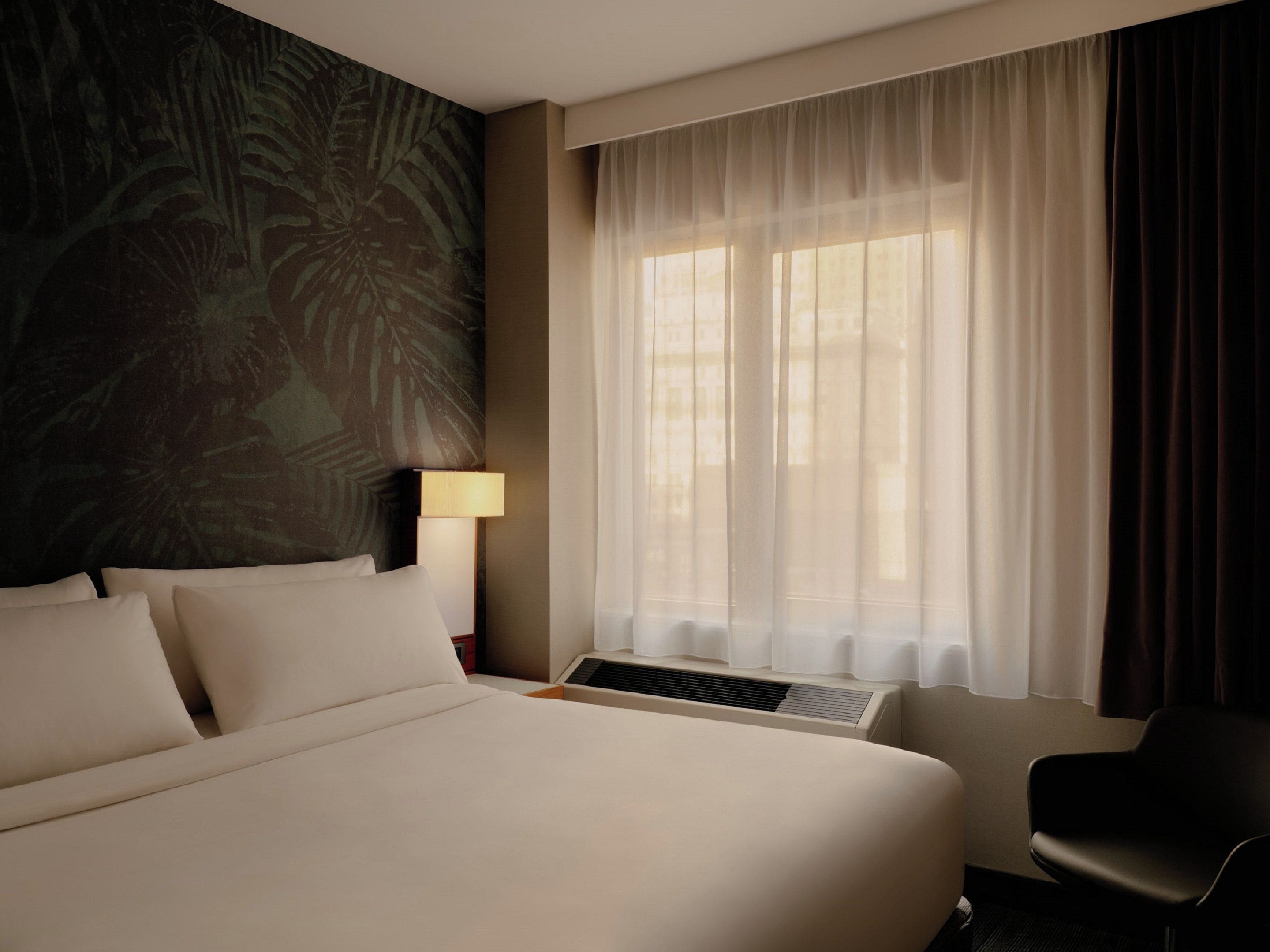 Habitación de hotel con una cama grande, un escritorio, una silla, una ventana con cortinas translúcidas y un diseño de hojas tropicales en la pared.