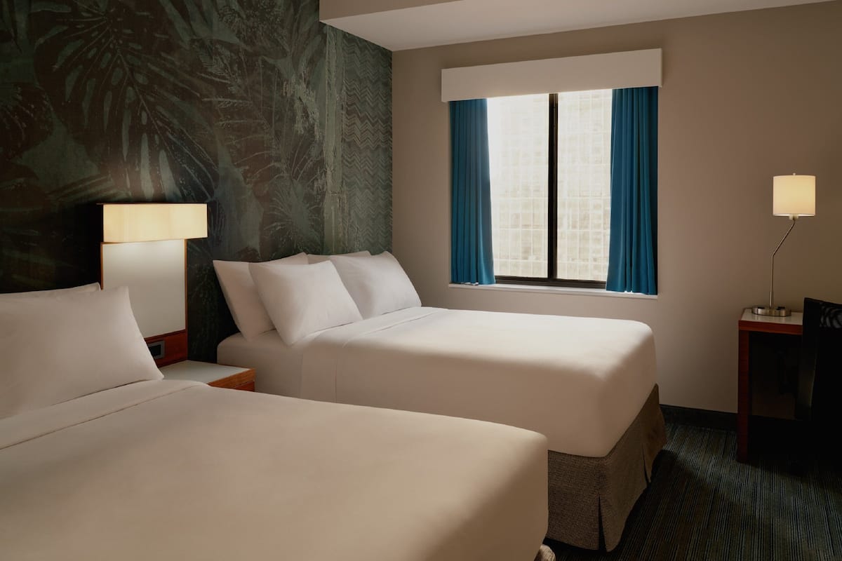 A hotel room with two beds, a window with curtains, a lamp, and a wall with a tropical mural.