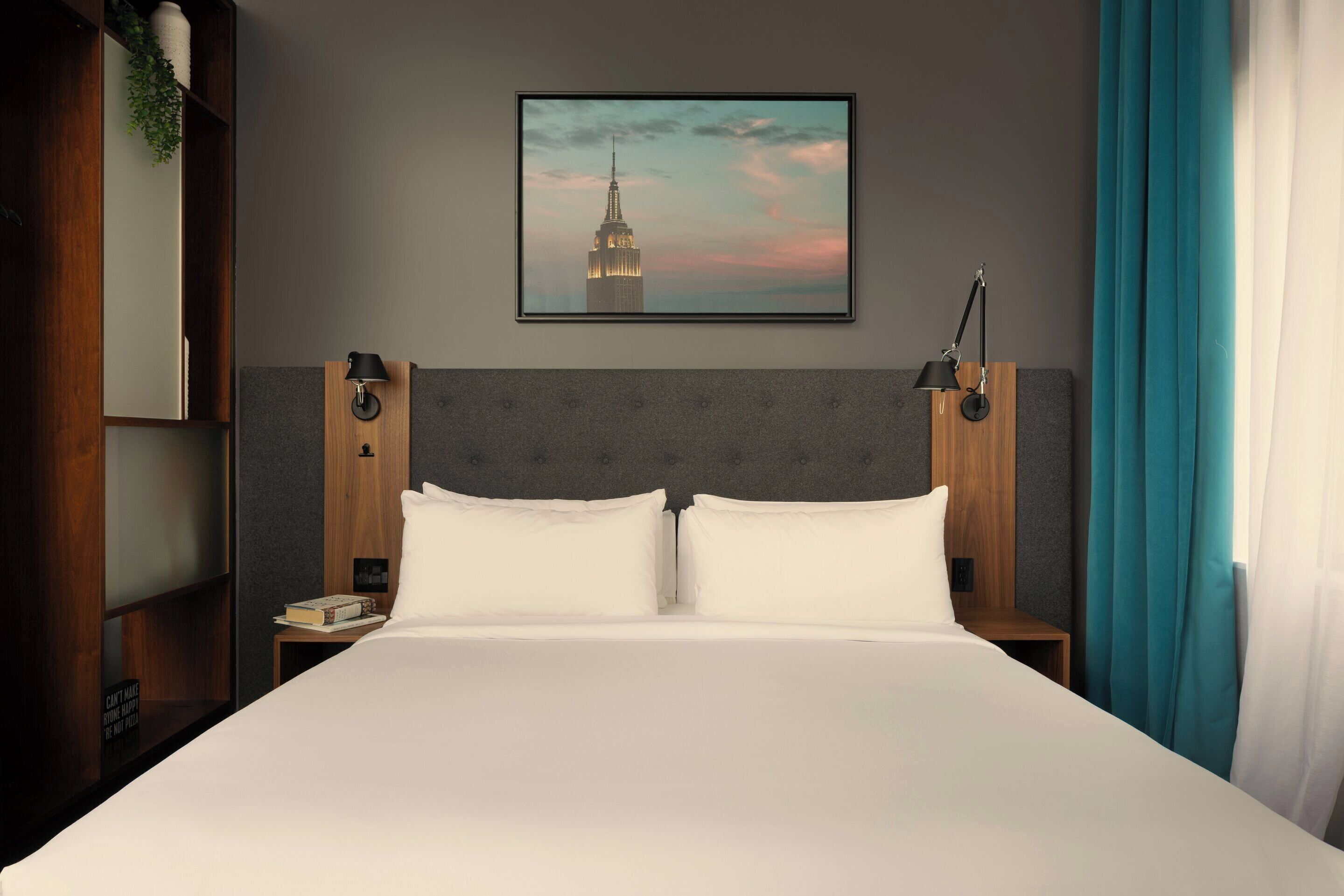 A hotel room with a large bed, two bedside tables, a nightstand, a framed picture of a building, and a window with curtains.
