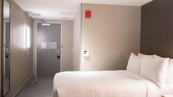 A hotel room with a bed, a door, a wall-mounted light, and a small red emergency light.