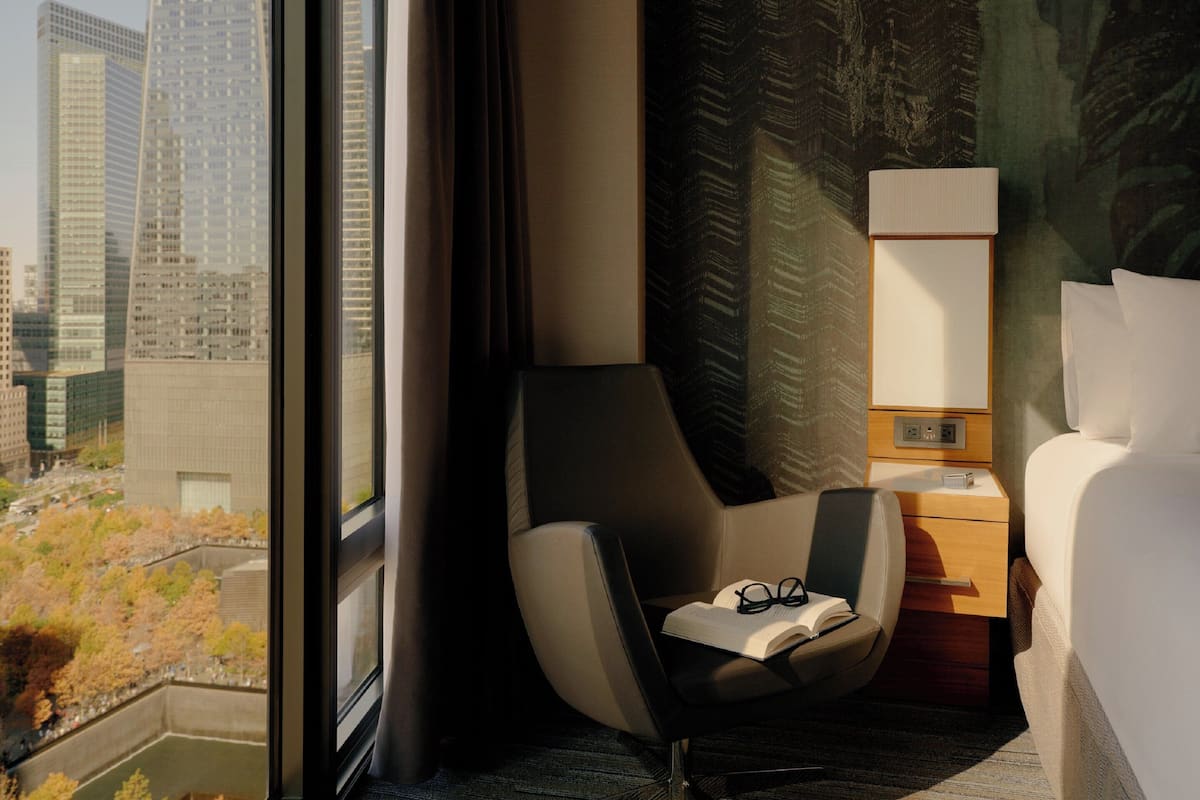 A hotel room with a view of tall buildings, a modern chair, a bedside table with a lamp, and a neatly made bed.