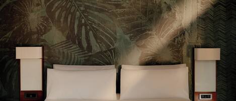 A bed with two pillows, flanked by two bedside tables with lamps, against a wall with a tropical leaf pattern.