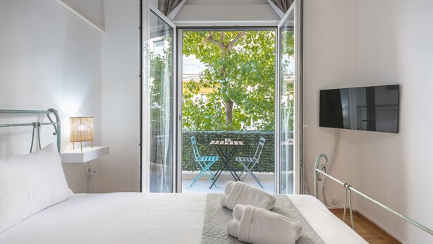 A hotel room with a bed, a TV, a balcony with a table and chairs, and a view of trees and buildings. - Athens Bright Suite by Cloudkeys (Athens)