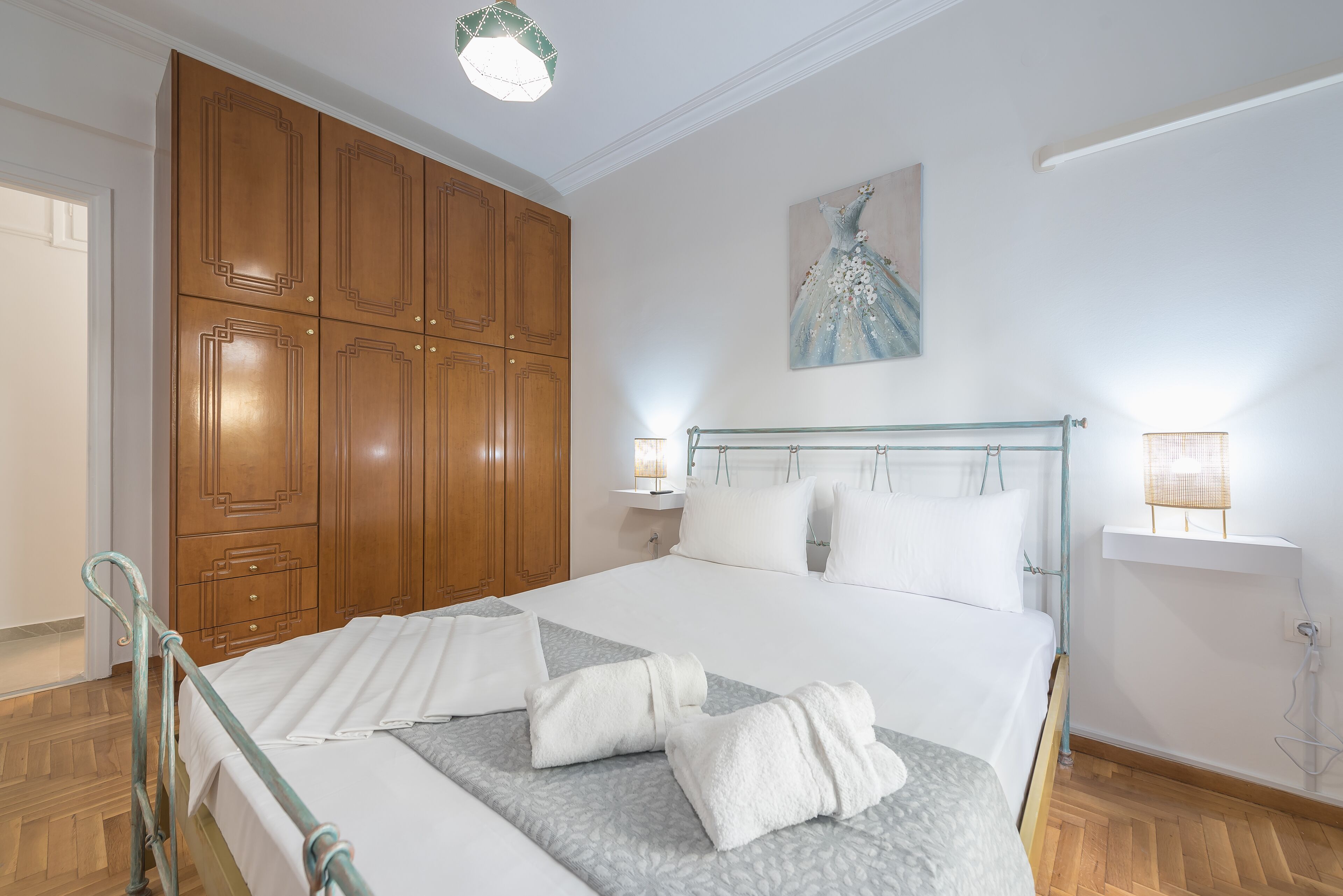Un lit bien fait, avec du linge de lit et des serviettes blanches, une armoire en bois, un tableau au mur et des lampes de chevet.