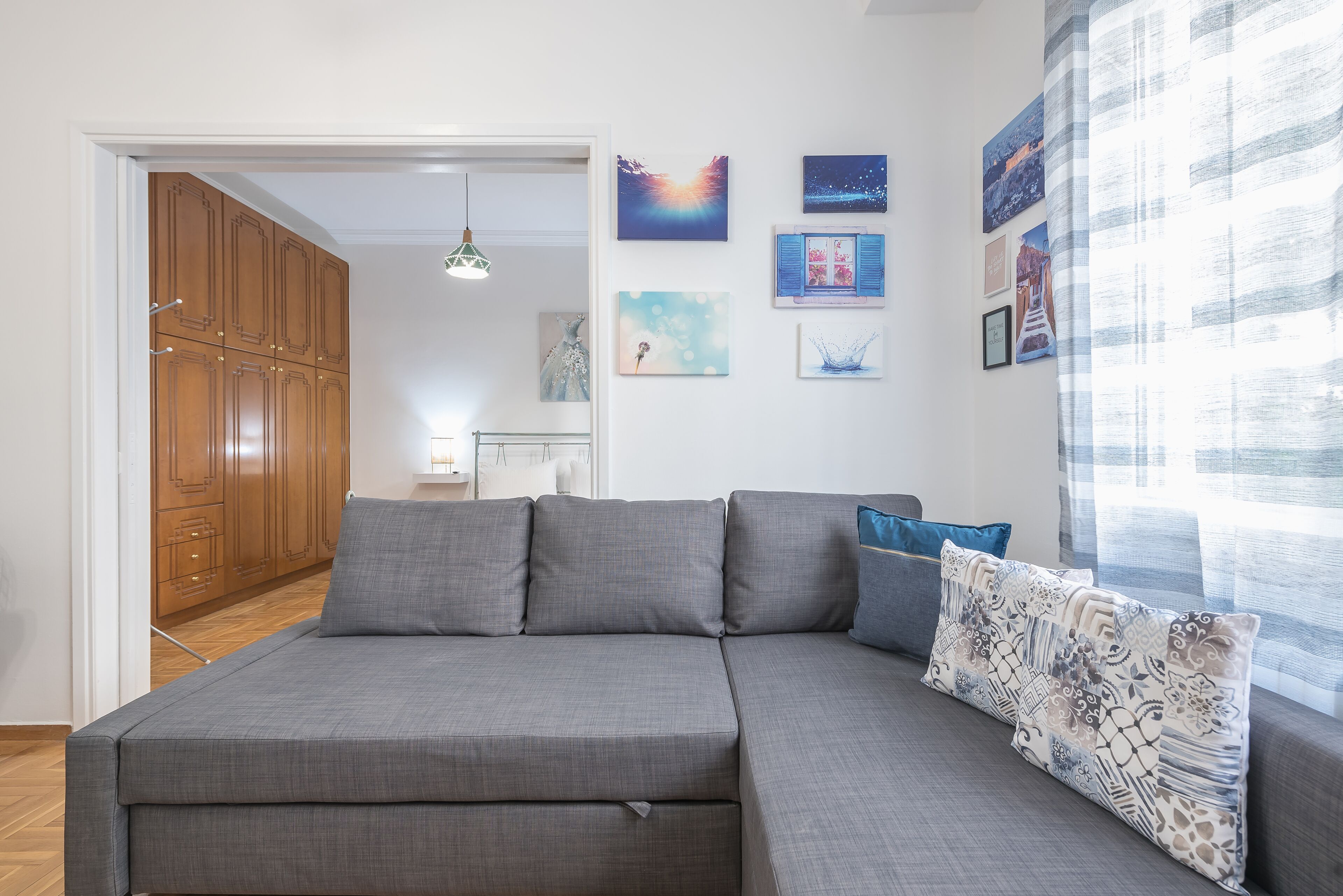 A modern living room with a grey sectional sofa, a patterned throw pillow, and a view of a wooden wardrobe and a dining area in the background.