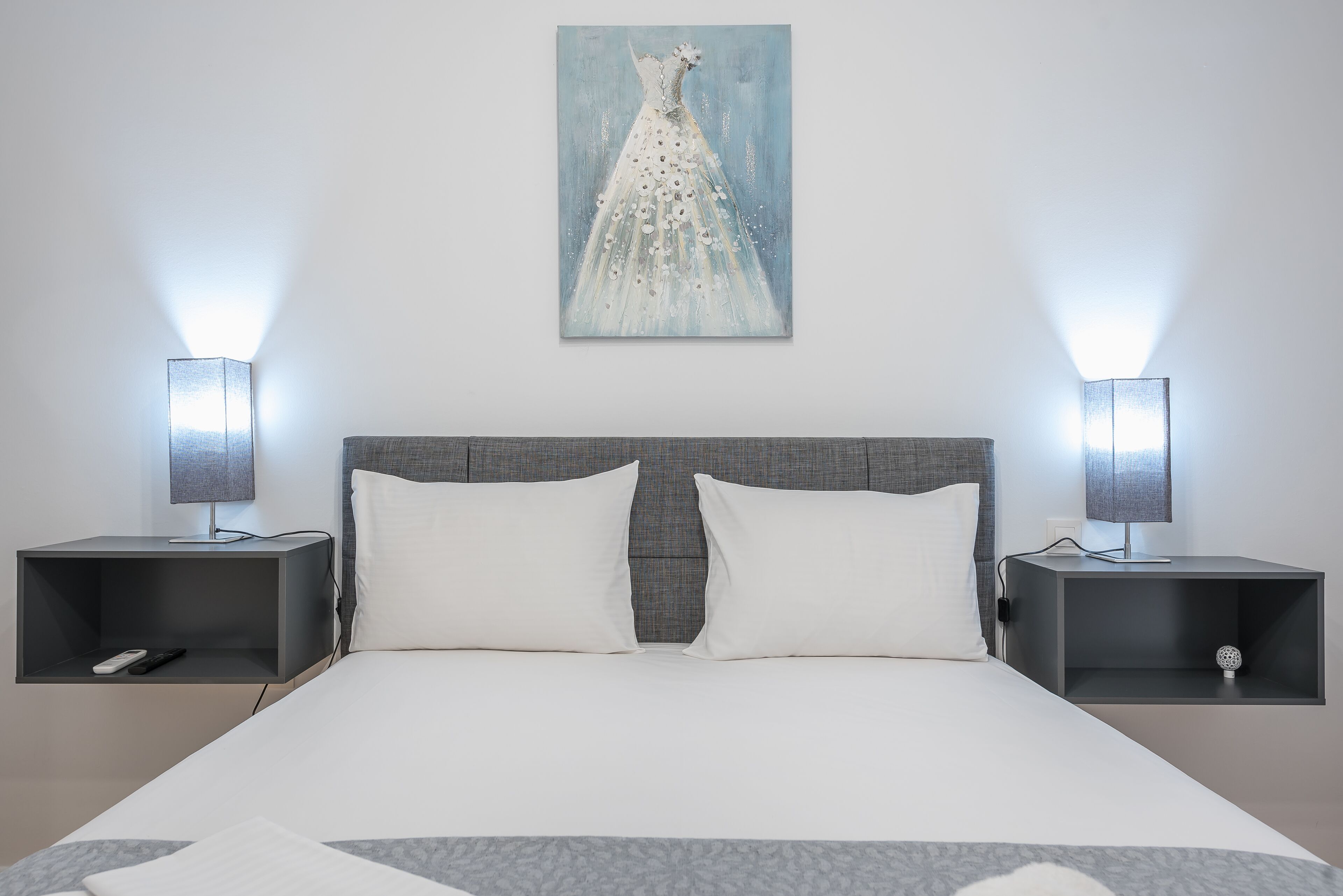 A modern hotel room with a large bed, two bedside tables with lamps, a wall-mounted painting of a dress, and a grey headboard.