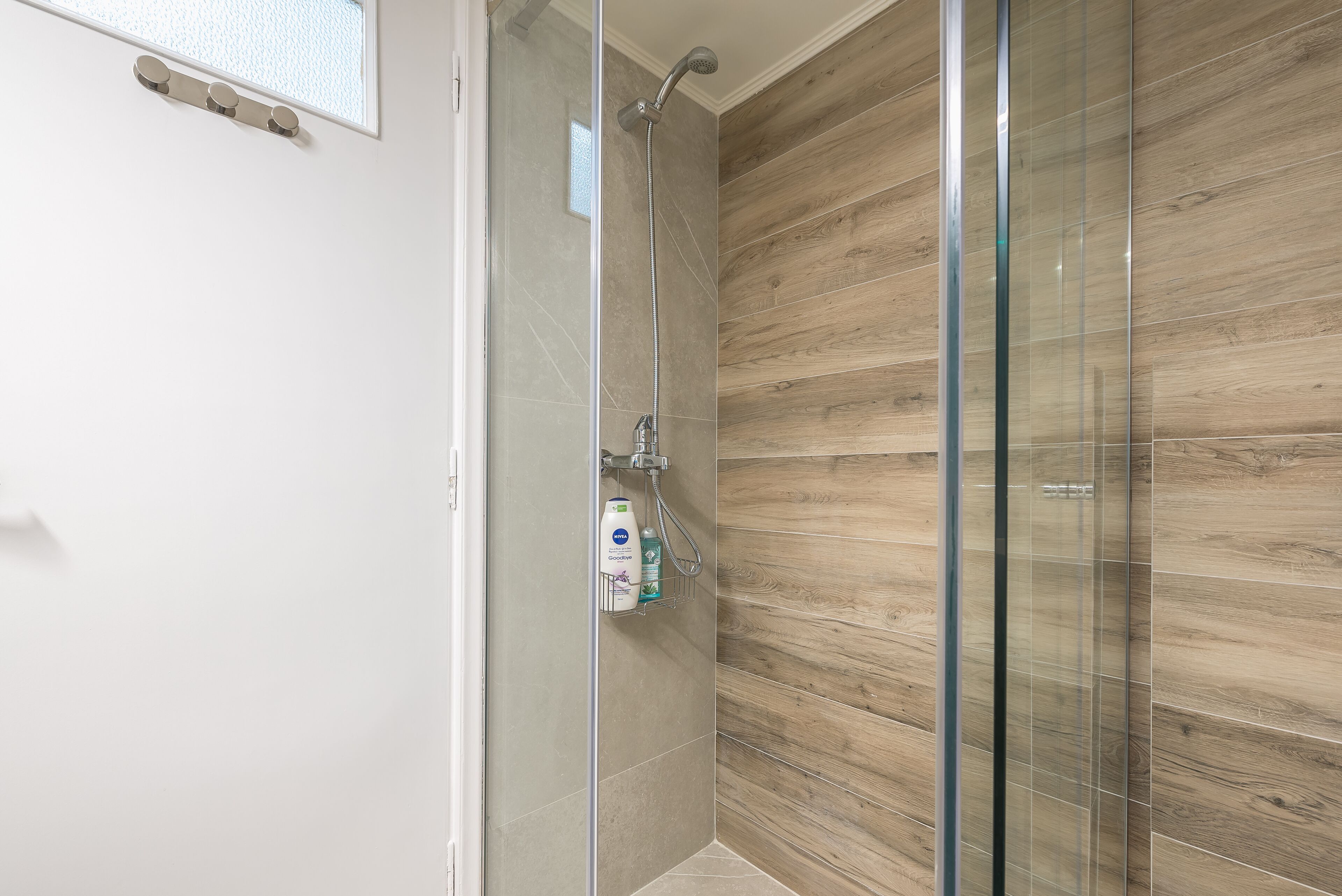 A modern shower area with a glass door, a wooden wall panel, and a shelf with toiletries.