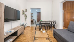 A modern living room with a flat-screen TV, wooden flooring, a grey sofa, a dining area with a table and chairs, and a wooden wardrobe. - Athens Bright Suite by Cloudkeys (Athens)