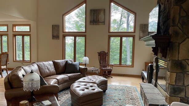 A spacious living room with a large brown leather sofa, a matching ottoman, a wooden coffee table, and a stone fireplace. There are two large windows providing natural light and views of the outdoors.