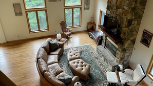 A spacious living room with a stone fireplace, large windows, a sectional sofa, and a television.