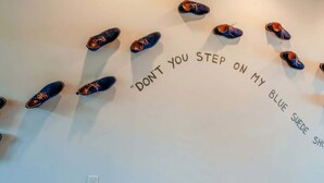Eine Wand mit mehreren Paar Schuhen, die in einer Halbkreisform angeordnet sind, und darüber der Text „DON'T YOU STEP ON MY BLUE SUDE SHOES“.