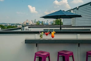 A rooftop bar with a city skyline view, three colorful cocktails, and purple stools.