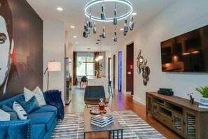 A modern living room with a blue sofa, a coffee table with books and a bottle, a wooden cabinet, and a large TV on the wall. - Fall Nashville getaway: 2 rooftop homes, karaoke rooms, sleeps 24 near Parthenon (Nashville)