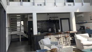 Een ruime woonkamer met hoge plafonds, een balkon met metalen hekwerk, een keuken met moderne apparatuur, en een zithoek met een blauwe tafel en witte kussens.