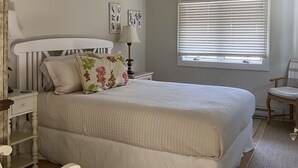 Een slaapkamer met een groot bed, een nachtkastje, een stoel, een raam met jaloezieën en een plafondventilator.