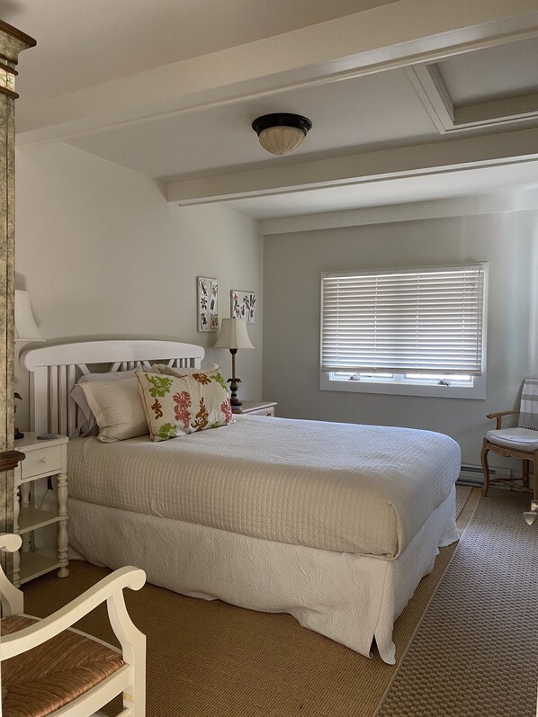 Une chambre à coucher avec un grand lit, une table de chevet, une chaise, une fenêtre avec des stores et un ventilateur de plafond.
