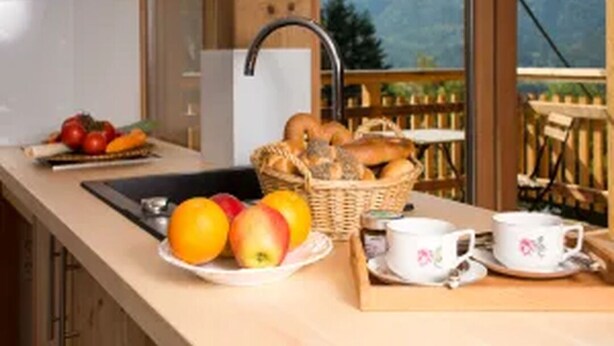 Een keuken met een spoelbak, een fruitschaal met appels en sinaasappels, een broodmand en een dienblad met kopjes en schotels.