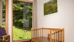 Een kamer met een houten bankje, een klein tafeltje met een paars kussen, en een groot raam met uitzicht op groen.