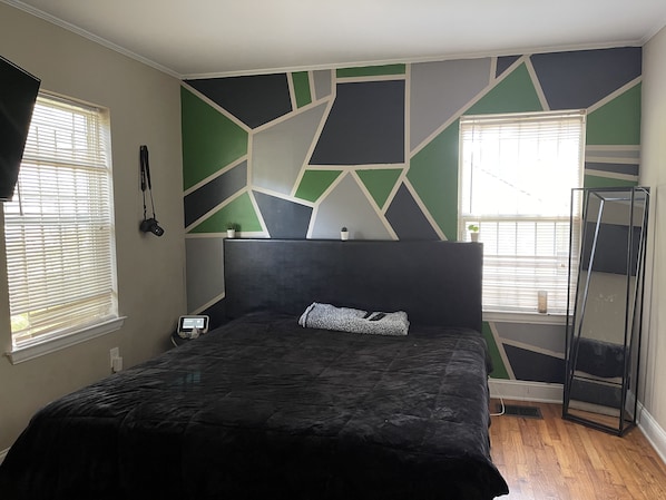A bedroom with a modern geometric wall design, a large bed with a dark headboard, and two windows with blinds.