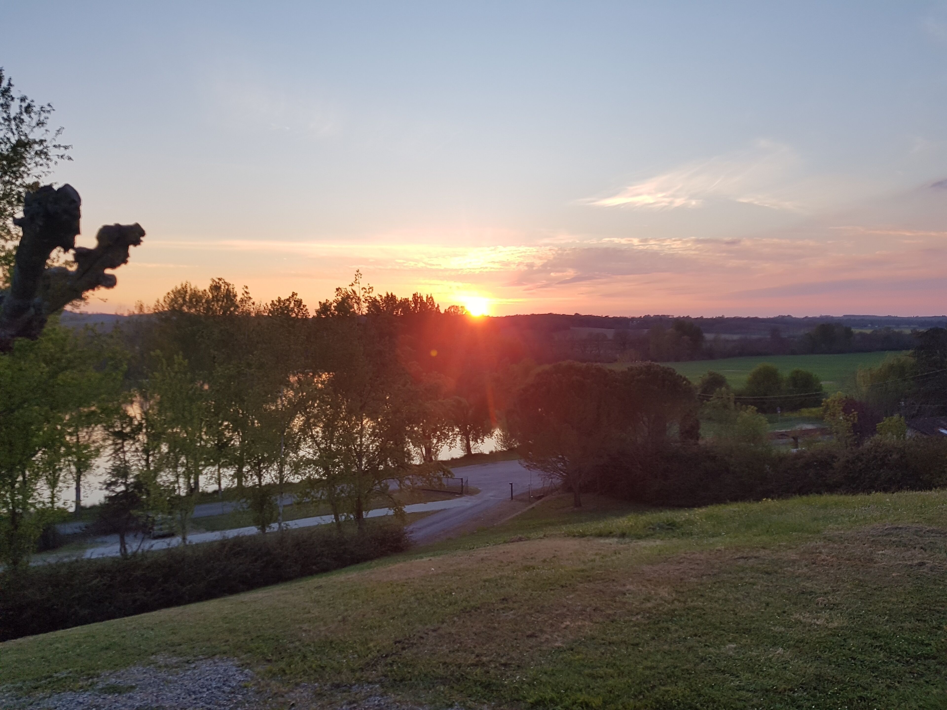 Een zonsondergang boven een landelijk landschap met water, bomen en een weg.