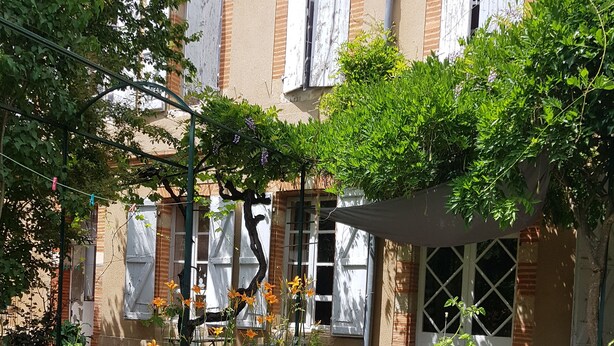 A courtyard with a variety of plants and flowers, including red flowers and orange lilies, in front of a building with white window shutters and a brick facade. - Large bedroom (3 + 1 baby ) & Dining area in a country village -  (Lézat-sur-Lèze)