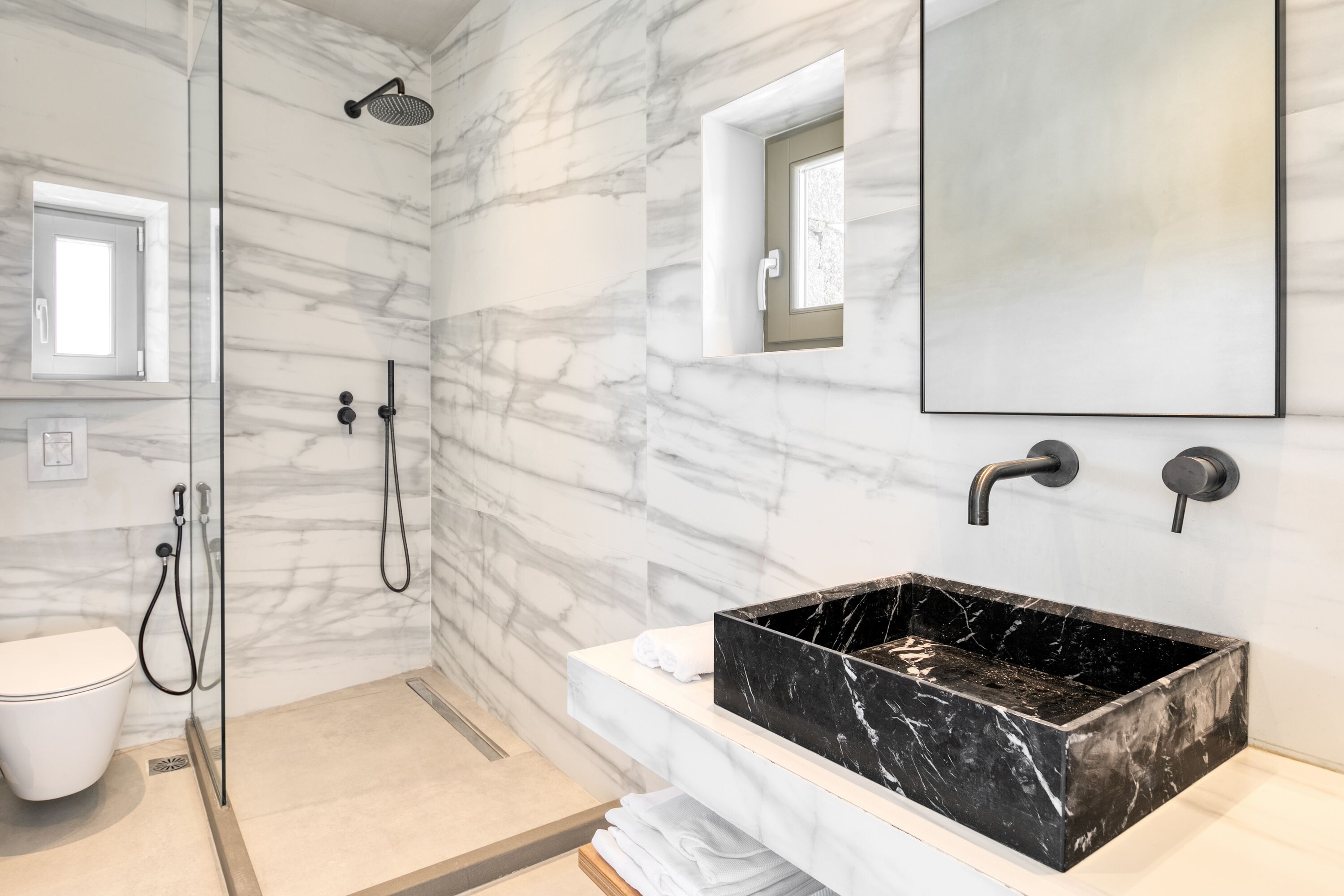 A modern bathroom with a marble wall, a black marble sink, a glass-enclosed shower, and a small window.