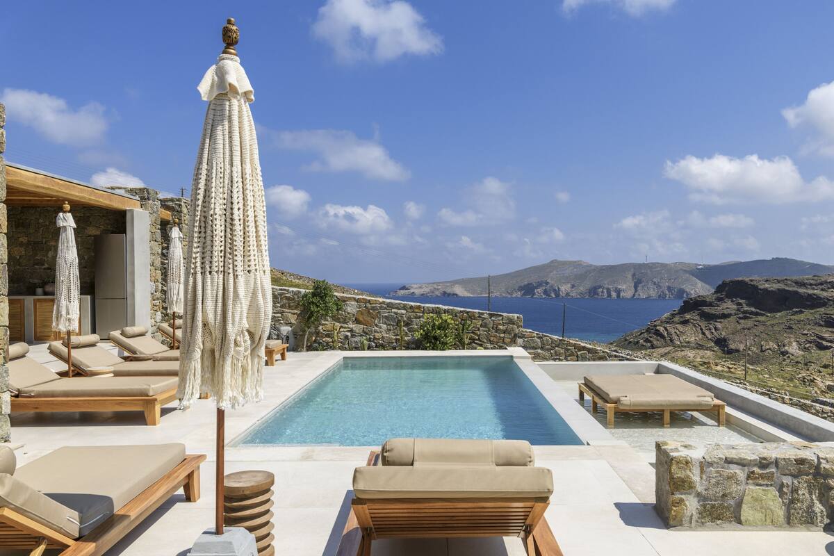 A poolside area with lounge chairs, a large umbrella, and a view of the sea and mountains.