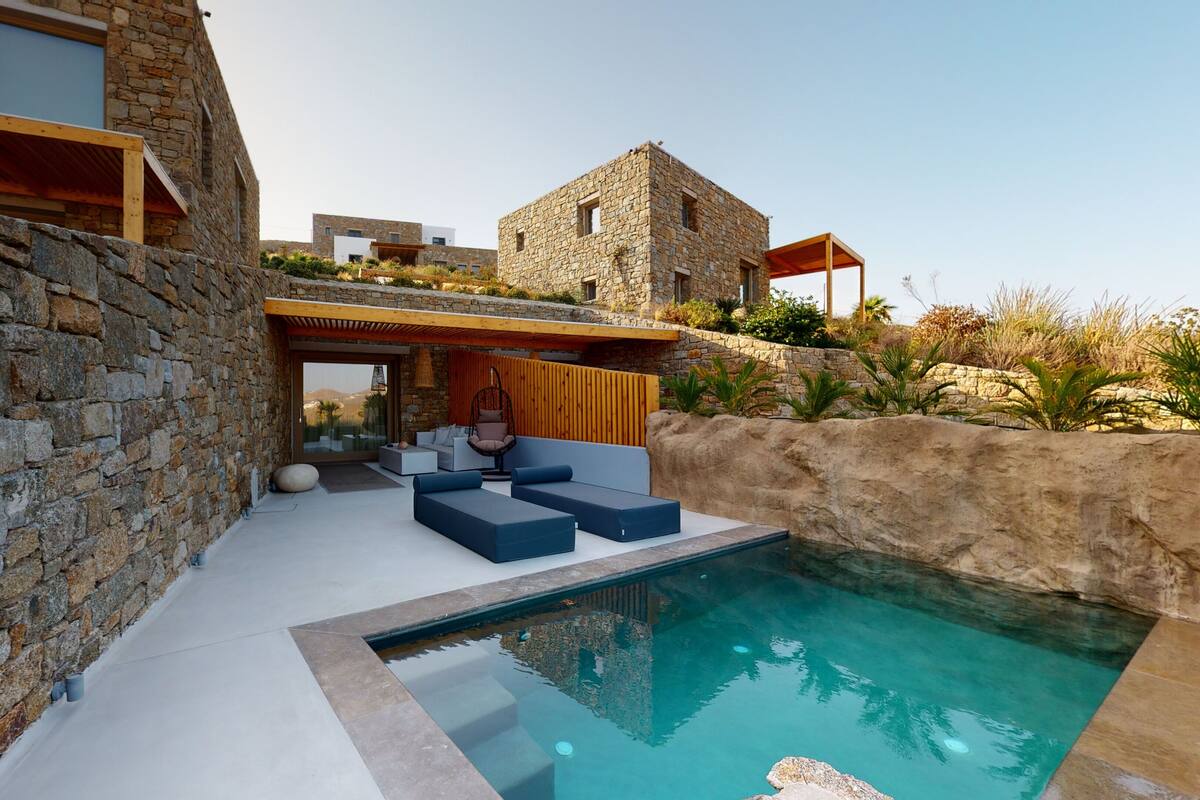 A modern outdoor pool area with stone walls, a hanging chair, and a seating area with cushions.