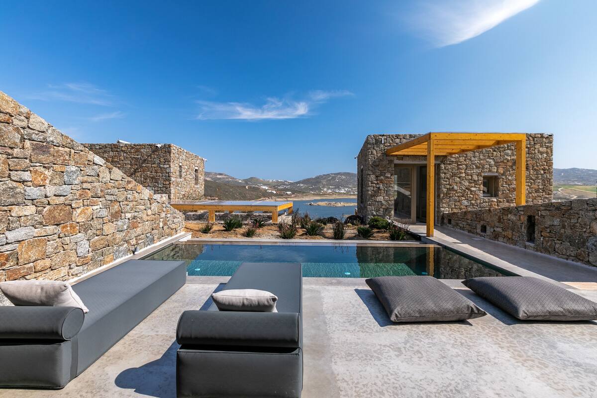 A rooftop pool area with stone walls, lounge chairs, and a view of the sea and mountains.