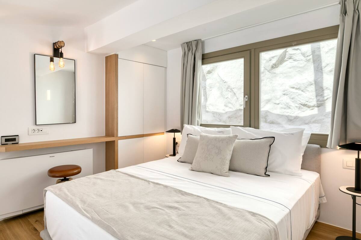 A modern bedroom with a large bed, a bedside table with a lamp, a mirror, and a window with curtains.
