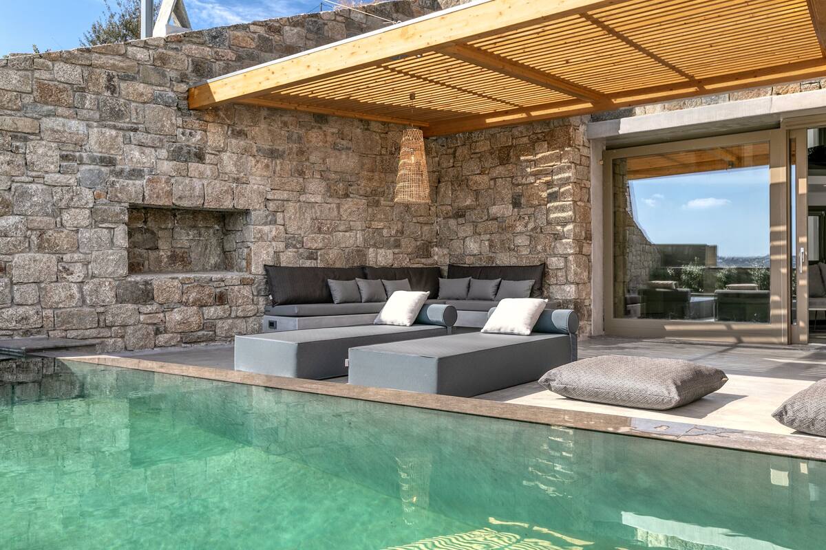 A poolside area with a stone wall, a wooden pergola, a modern sofa with cushions, and a clear pool.