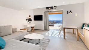 A modern living room with a sofa, coffee table, and a dining area with a table and chairs. A flat-screen TV is mounted on the wall, and there is a view of the outdoors through a large window. - Panormos Bay Suites (Mykonos)