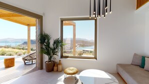 A modern living room with a large window offering a scenic view of a coastal landscape. - Panormos Bay Suites (Mykonos)