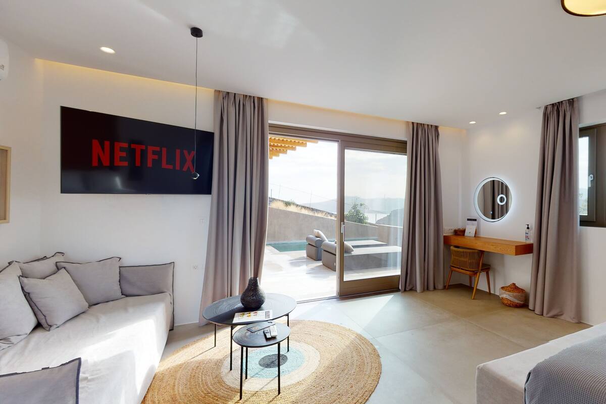 A modern hotel room with a large window, a sofa, a small table, a desk with a mirror, and a sign for Netflix.