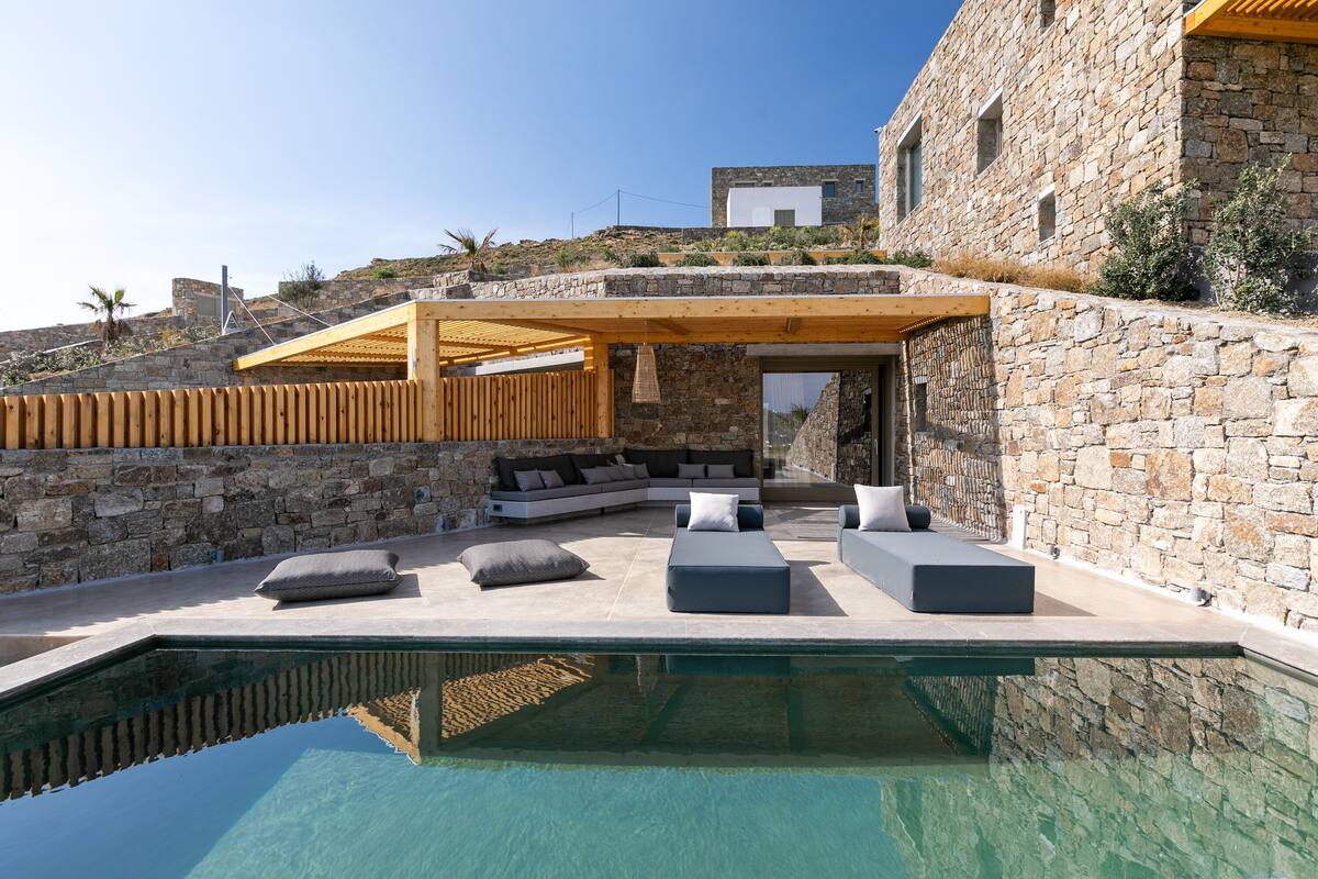 A modern outdoor pool area with stone walls and a wooden pergola, featuring a built-in seating area with cushions.
