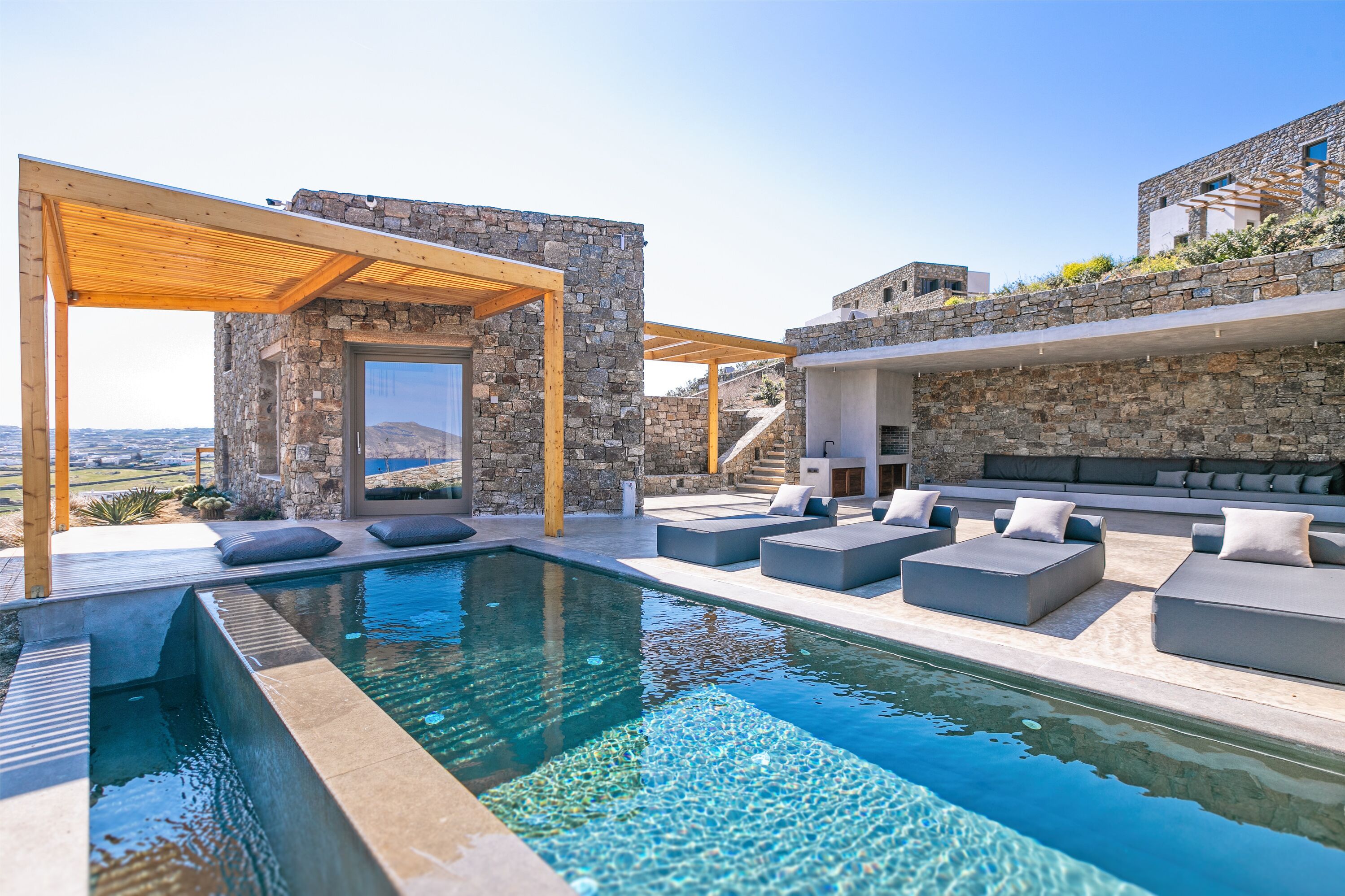 Un área de piscina moderna al aire libre con paredes de piedra, pérgolas de madera y asientos cómodos.